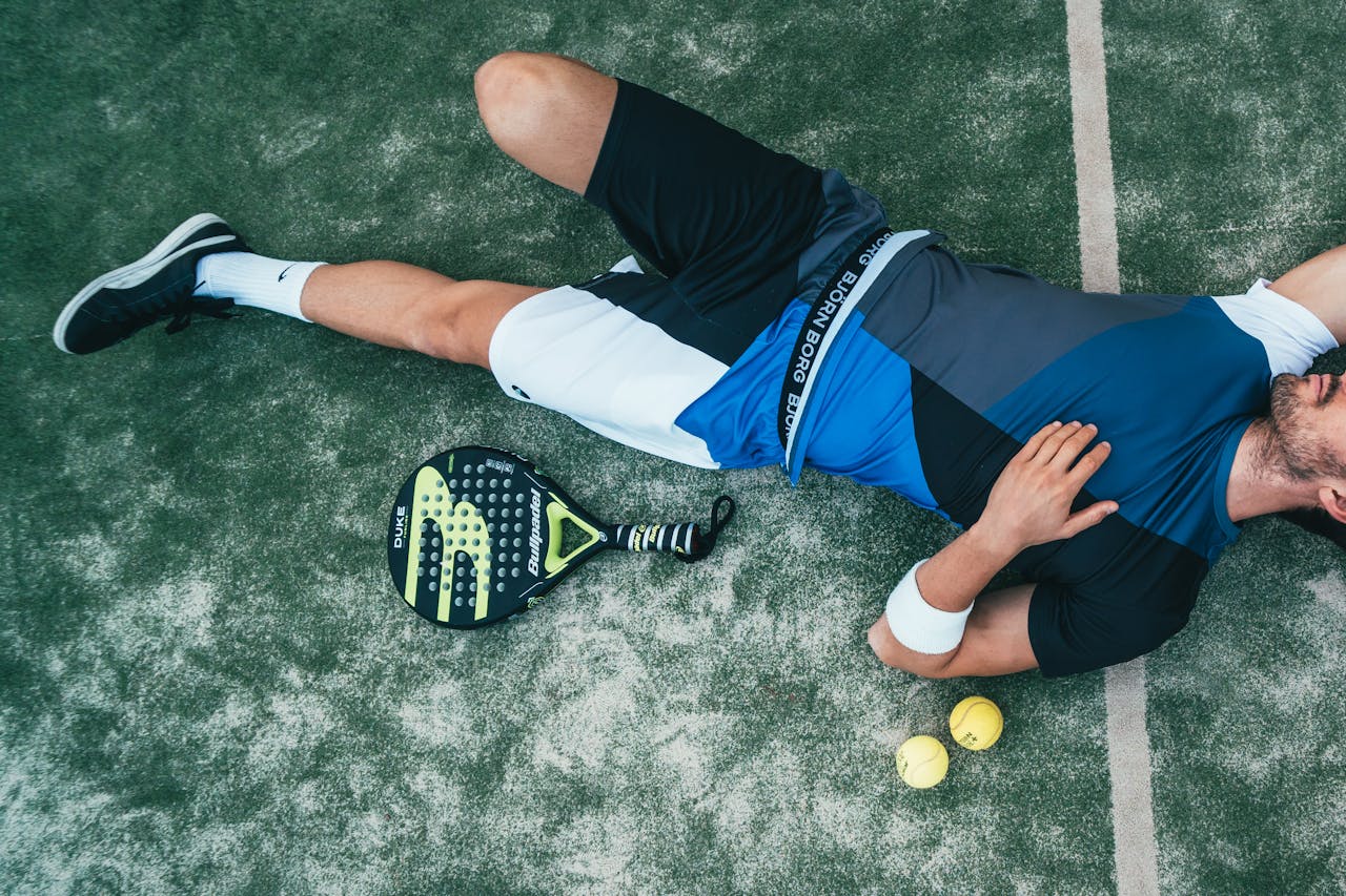 Tired young athlete resting on pádel court with racket and balls nearby.